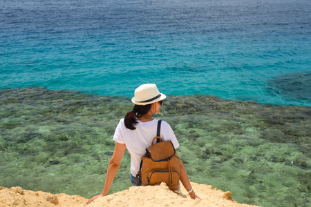 A young tourist with a backpack sits on the rocks by the sea and looks into the distance. Rest, relaxation, travel, vacation.の写真素材