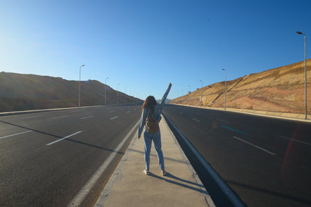 A girl in a denim suit with a backpack walks in the center of a quiet road among the mountains at...