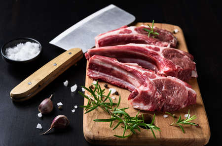 Two raw lamb chops on a wooden cutting board with spices on a dark wooden background