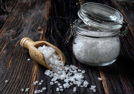 Sea salt in a wooden scoop and glass jar on wooden background. Coarse salt.