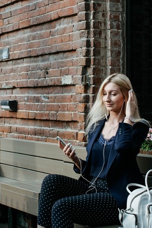 Young attractive woman listening to music on her smartphone while sitting on a street bench during a lunch breakの写真素材