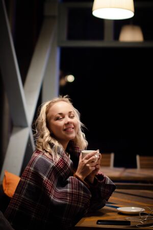 Image of a young beautiful blonde woman to a street cafe. The blonde drinks tea wrapped in a warm...