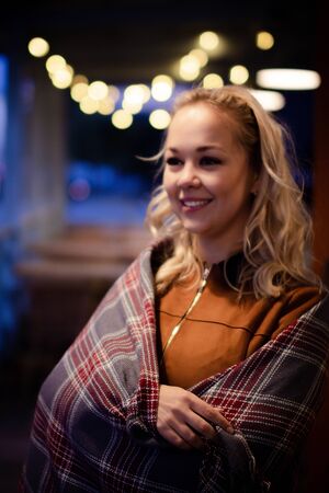 Young pretty blonde with a plaid on her shoulders on a cozy terrace on Christmas evening. Cozy autumn evening.の写真素材