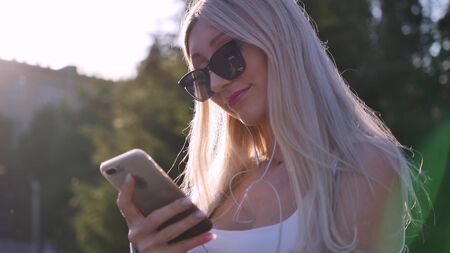 Attractive young woman listening to music in headphone use smartphone at city walk sunset look around smile portrait close up.の写真素材
