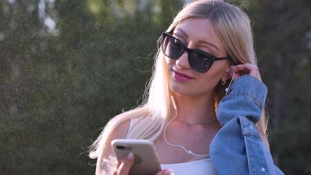 Attractive young woman listening to music in headphone use smartphone at city walk sunset look around smile portrait close up.の写真素材