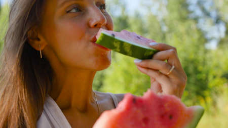 Close-up of an attractive cute young woman with kind eyes eating a piece of watermelon and smiling at the camera. A beautiful and pretty girl is enjoying the taste of fruit outdoors in the park.の写真素材