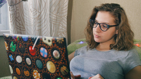 young adult woman sitting in the living room on the couch using a laptop. A woman with glasses works or studies from home on the Internet on a computerの写真素材