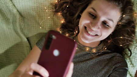 A young charming woman with lights in her hair takes a selfie on her smartphone. Romantic atmospheric photo about dreams. Winter Holidays Concept.の写真素材