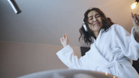 beautiful asian woman in white robe listens to music with wireless headphones in the bathroom.