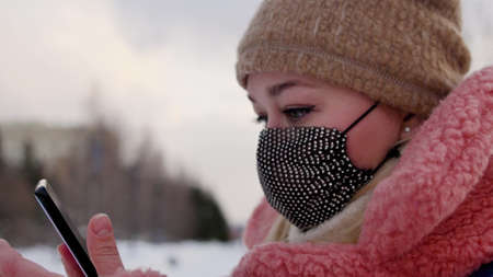 Portrait of a beautiful young girl in winter clothes with a mask on her face who is talking on the phone outdoorsの写真素材