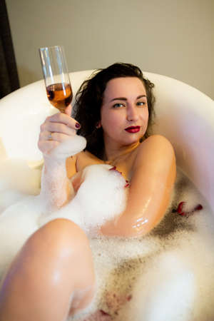 Pretty brunette takes a bubble bath. Woman holding a glass of champagne while lying in the bathroomの写真素材