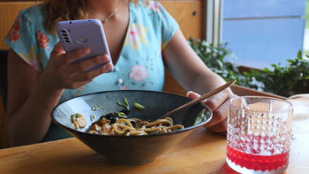 A woman is watching the news on her smartphone in an Asian restaurant. Woman eating noodles with vegetables and seafood with sushi chopsticks. Delicious udon.の写真素材
