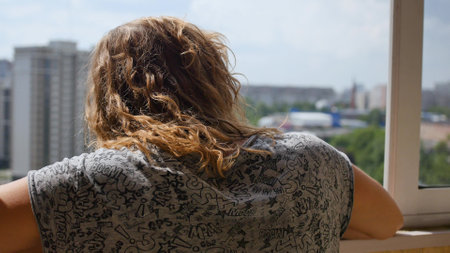 Cute curly-haired woman opens the window to breathe fresh air, Woman looks out the window at the city in the morning sunの写真素材