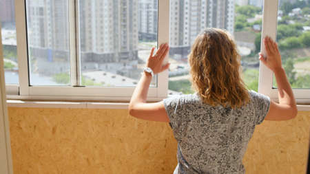 Cute curly-haired woman opens the window to breathe fresh air, Woman looks out the window at the city in the morning sunの写真素材