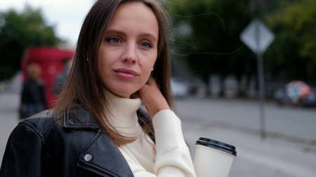 Happy caucasian business woman in a white sweater and black leather jacket walks down the street to her company office holding coffee in her handsの写真素材