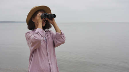 Beautiful asian woman in a hat stands on the seashore and looks through binoculars and enjoys a beautiful dayの写真素材