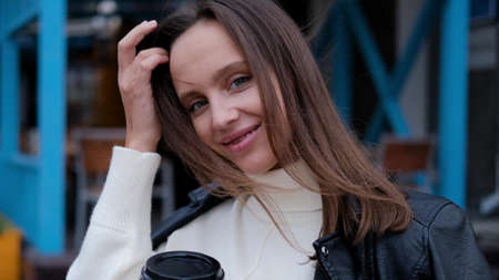 Happy caucasian business woman in a white sweater and black leather jacket walks down the street to her company office holding coffee in her handsの写真素材