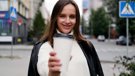 Happy caucasian business woman in a white sweater and black leather jacket walks down the street to her company office holding coffee in her handsの写真素材
