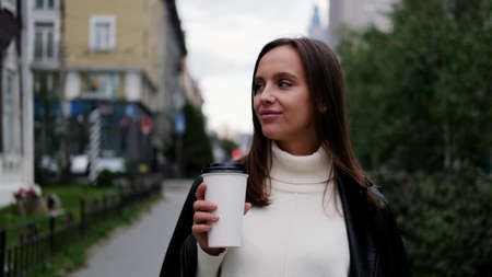 Happy caucasian business woman in a white sweater and black leather jacket walks down the street to her company office holding coffee in her handsの写真素材