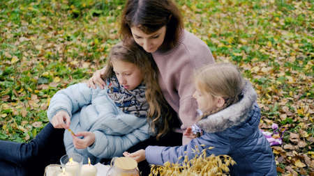 young mother and two little daughters are drinking tea at a picnic in nature.