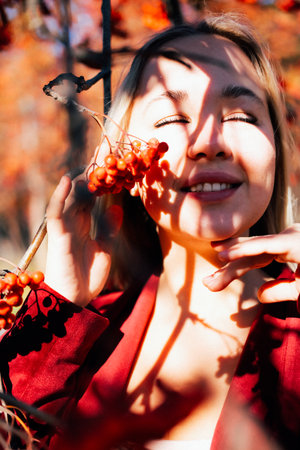 Young beautiful blond woman in the rays of the sun on a background of rowan branches in autumn. Autumn atmosphere and moodの写真素材