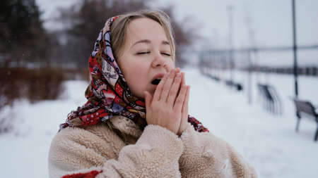 Young beautiful woman in a fur hat warms her hands on the background of a winter snow-covered park.の写真素材