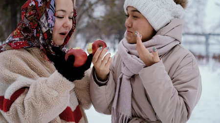 Young female friends walk in a winter park and bite ripe red apples. Organic food.の写真素材