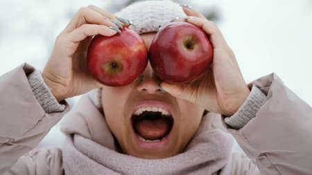 crazy joyful woman makes faces with two apples in her hands against the background of a snowy winter parkの写真素材