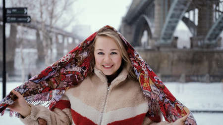 A young girl in a beautiful bright headscarf poses at the camera against the background of a snow-covered city embankment.の写真素材