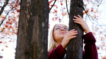 Cute blonde hugs a tree in the autumn city park. Beautiful woman gently hugs a tree trunk in the fall season.の写真素材