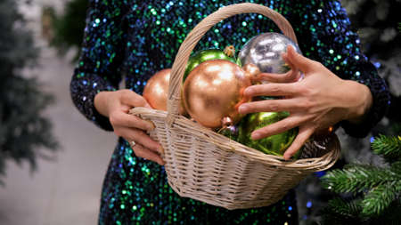 A woman in a shiny dress holds a basket with Christmas balls in her hands against a background of...