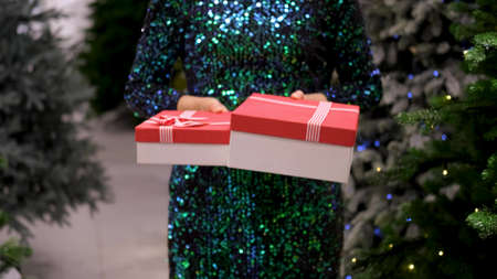 Womens hands are holding out gifts in festive packaging. Happy woman in a bright shiny dress gives...