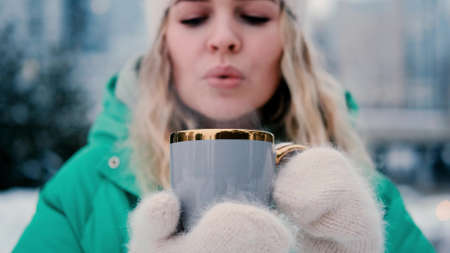 A cute caucasian girl in warm clothes drinks hot tea from a mug while walking in a snowy city park.の写真素材