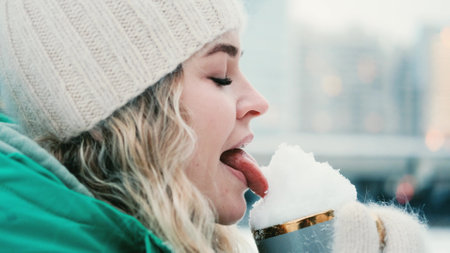 The girl licks the snow in a mug on the background of a winter park.の写真素材