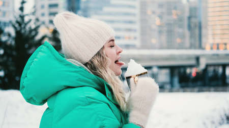 The girl licks the snow in a mug on the background of a winter park.の写真素材