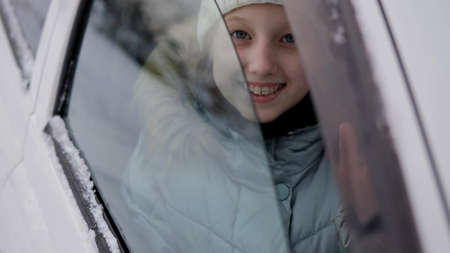 A child in warm winter clothes looks and smiles at the camera through the car window. Winter portrait of a little girl in a carの写真素材