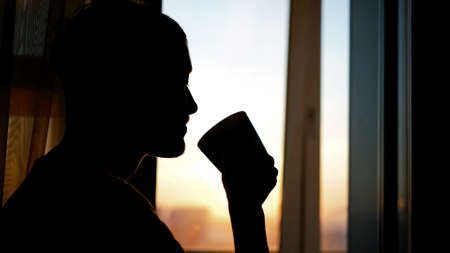 A sad, depressed man is drinking tea at the window of his apartment during a beautiful sunset in the winter sun.の写真素材