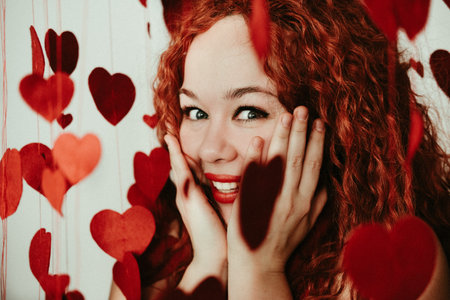 Carefree girl with long red hair expressing positive emotions on valentines day. Excited lady looks at red hearts and laughs.の写真素材