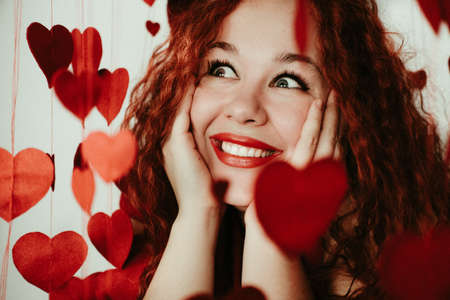 Carefree girl with long red hair expressing positive emotions on valentines day. Excited lady looks at red hearts and laughs.の写真素材