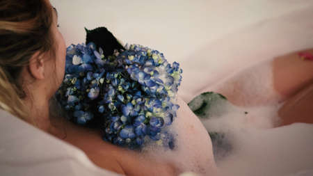 Close-up of a relaxed woman relaxing in a bubble bath. A woman touches her skin with a blue hydrangea flower.の写真素材