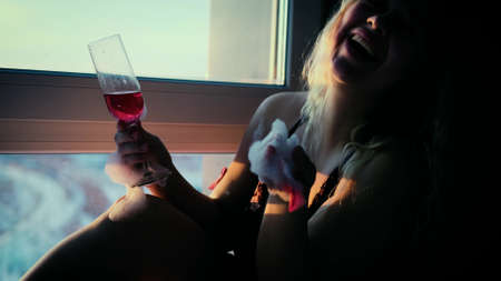 A very beautiful blonde sits on the edge of a bathtub filled with foam and drinks champagne. Relaxed woman enjoying the setting sun in her bathroomの写真素材