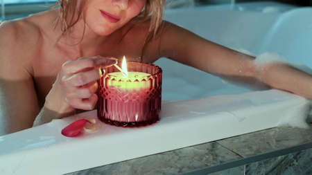 Relaxed woman lighting a candle while taking a bubble bath in the evening.の写真素材