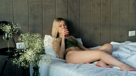 A woman in beautiful lingerie drinks wine while lying in bed. Relax at homeの写真素材