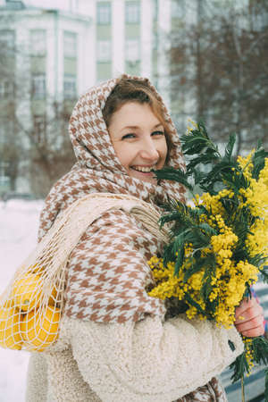 A happy woman rejoices at the arrival of the long-awaited spring. Woman in a snowy city with a bouquet of spring flowers. Yellow mimosa flowers are a symbol of spring and International Womens Dayの写真素材
