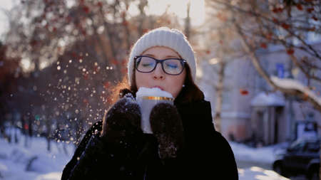 A young woman blows snow off a coffee mug on a snowy city street on a winter dayの写真素材