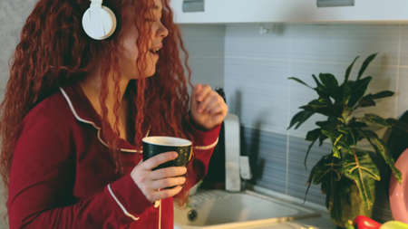 Portrait of a carefree curly woman who cooks in the kitchen and sings. Pretty cheerful red-haired...