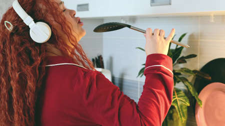 Portrait of a carefree curly woman who cooks in the kitchen and sings. Pretty cheerful red-haired woman listens to music on headphones in the kitchenの写真素材
