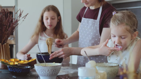 Two girls and mother are preparing cupcakes in the kitchen. Little daughter eats cream