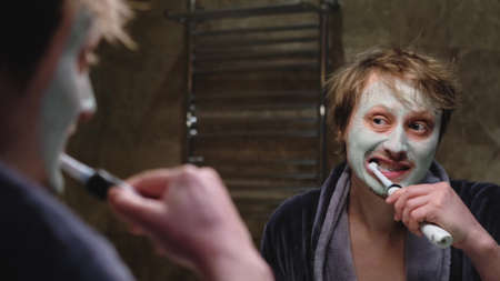 A man in a bathrobe brushes his teeth while looking in the mirror in the bathroom. The man applied a cosmetic mask to his face. Cosmetic procedures for mens skin care.の写真素材