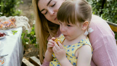 A young woman and little daughter are sitting in the backyard at a table with ripe organic berries and pastries. Summer holiday conceptの写真素材